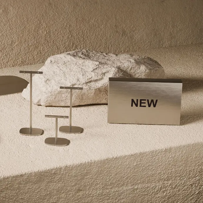 Three small T-shaped brushed metal display stands in different heights next to a pale rock and a brushed metal block with the word “NEW”, on a textured sand-coloured surface.